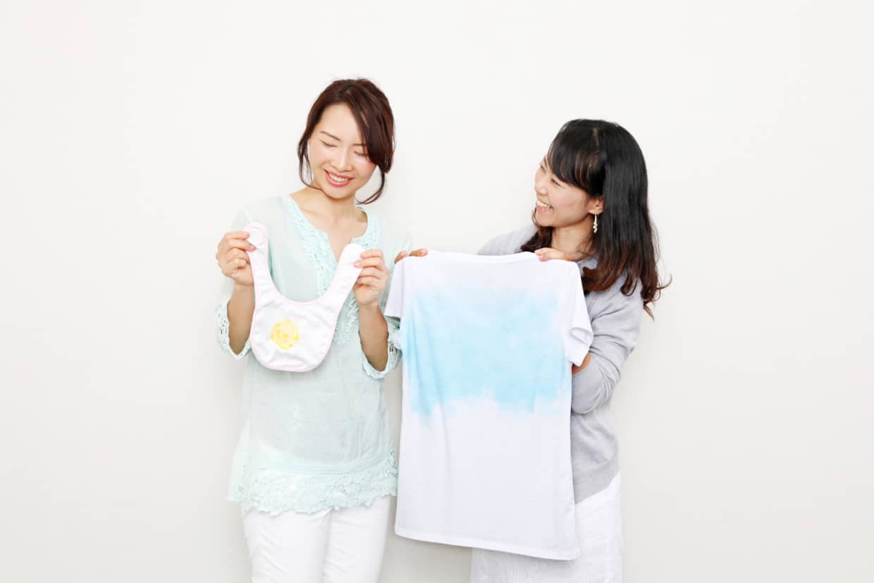 離乳食の食べこぼしのついたスタイ、汗を模した色水をかけたTシャツを持つ2人。
